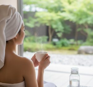 岩盤浴で休憩している女性（後ろ姿）が、手元にある温かいお茶のカップに手を添え、リラックスしつつ水分補給も意識している画像
